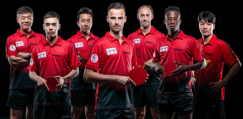 Trotz der leichten Abdunklung schimmern die roten Trikots des TTC Neu-Ulm auf dem Mannschaftsfoto ganz gut durch. Das Team von Trainer Chen Zhibin (l.) ist prominent besetzt. Neben dem Coach stehen Kay Stumper, Gustavo Tsuboi, Tiago Apolonia, Viktor Brodd, Abdel-Kader Salifou und An Jaehuyn (v.l.n.r.). (©Atelier Schlieper)
